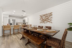 A modern kitchen with white cabinetry, a dining area with a wooden table and woven chairs, and a decorative wall piece. - Heated Pool • Home Gym • Walk to Beach (Holmes Beach)