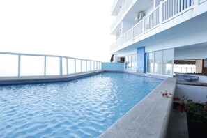 A swimming pool with a clear view of the sky, a white balcony with railings, and a section of the building's exterior wall.