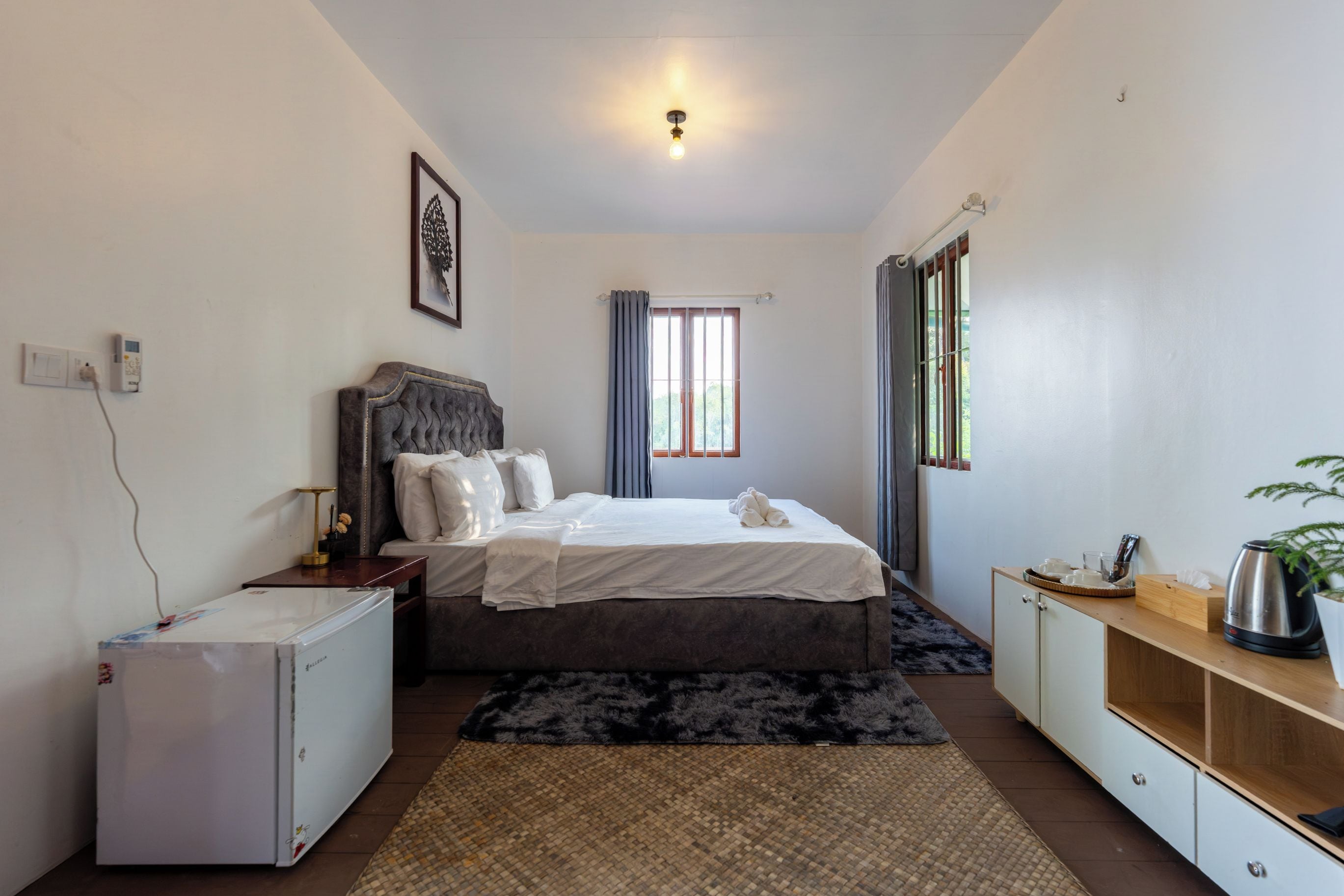 A bedroom with a bed, bedside table, a small refrigerator, a window with curtains, and a plant on a wooden cabinet.