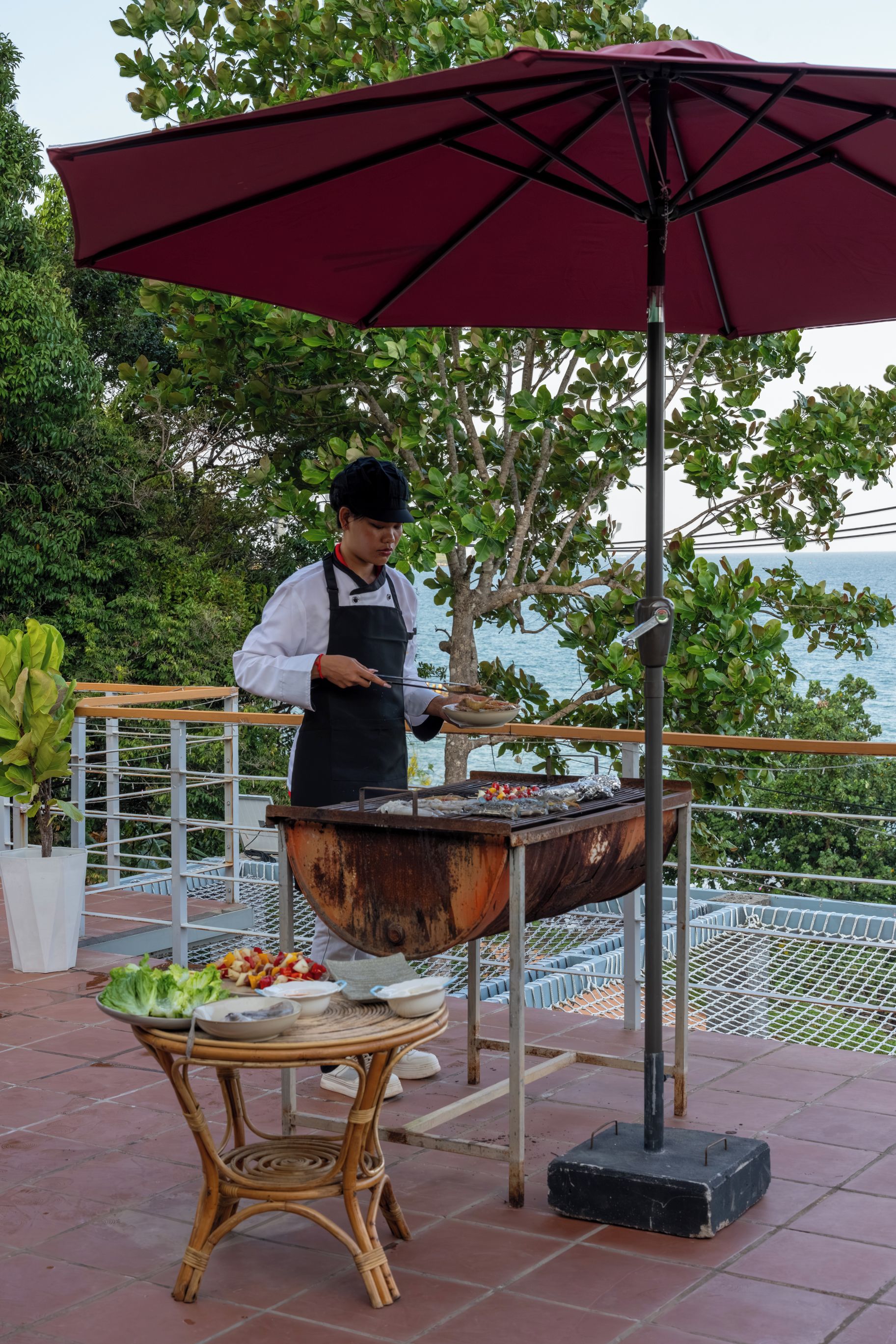 A chef grilling outdoors with a table of food and a red umbrella.