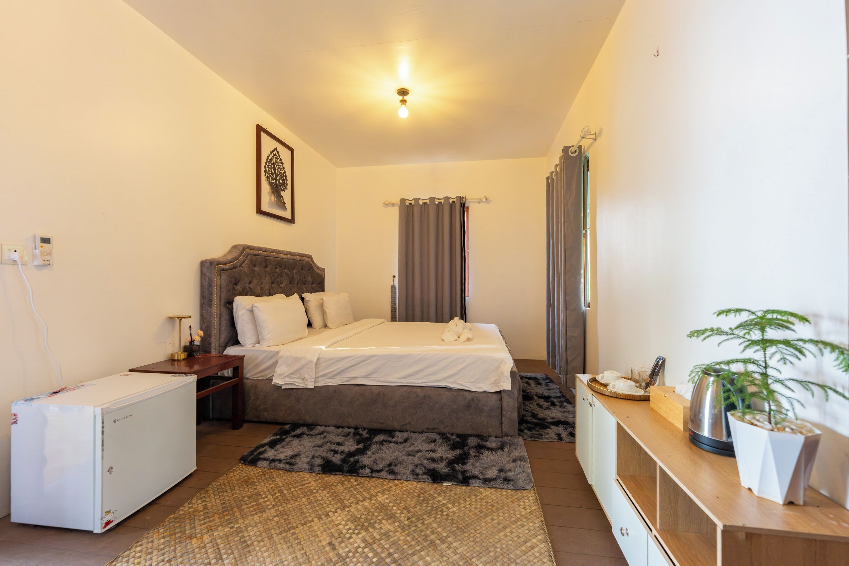 A bedroom with a bed, bedside table, a small refrigerator, a desk with a plant, and a window with curtains.