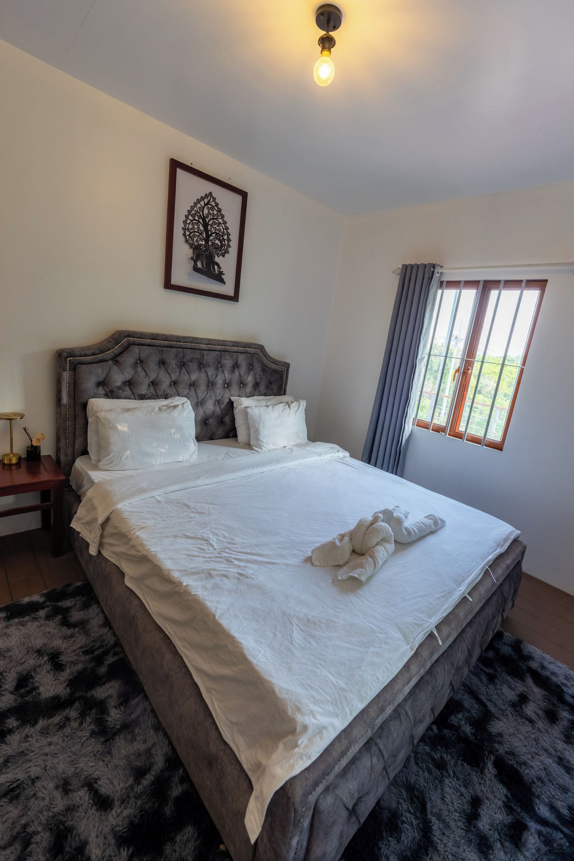 A bedroom with a large bed, a bedside table, a wall-mounted light fixture, a framed artwork, and a window with curtains.
