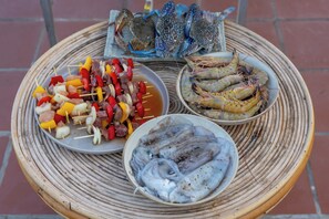 A round woven table with three dishes: skewers of meat and vegetables, a plate of shrimp, and a plate of fish.