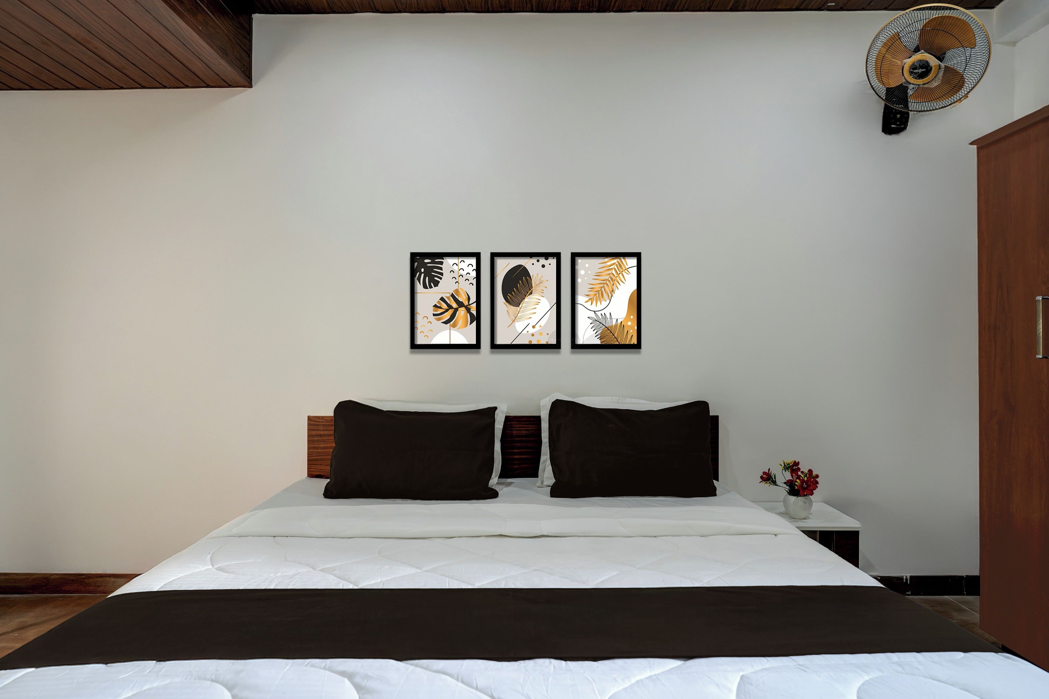 Un dormitorio con cama, dos almohadas, una mesita de noche, un florero con flores y tres cuadros en la pared.