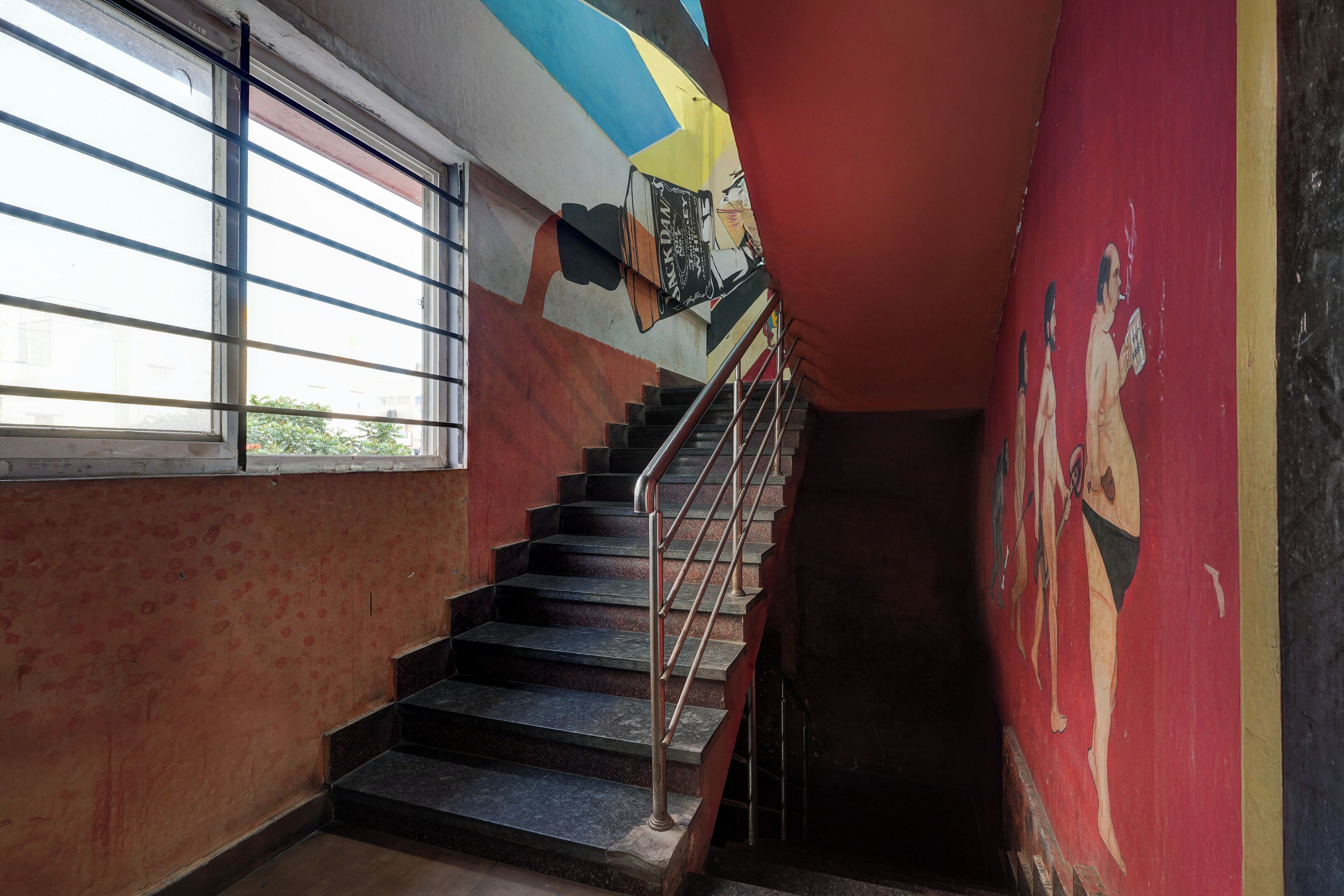 Una escalera con barandales de metal que baja a un nivel inferior, una ventana a la izquierda y una pared con un mural a la derecha.