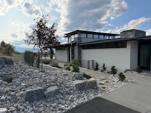 A modern house with a stone facade, a large window, and a well-maintained garden with rocks and small trees. - New luxury 4 bedroom/4 bathroom home on vineyard with pool and panoramic views. (Kelowna)