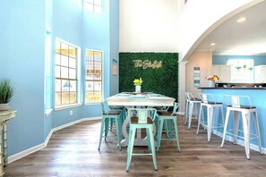 A dining area with a table and chairs, a green wall with a sign, and a kitchen in the background. - Steps to the Sand - 14 Beds - Sleeps 19 + Hot Tub (Surfside Beach)