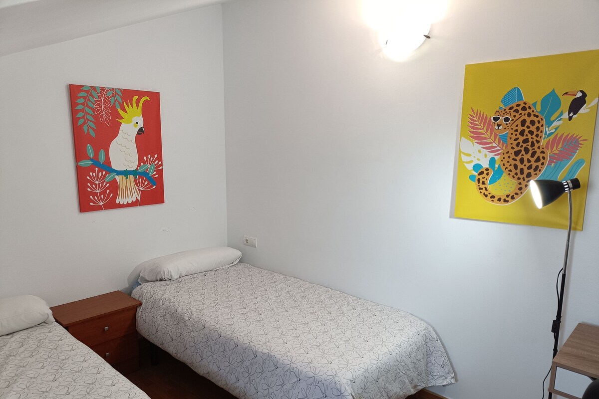 A room with two single beds, a wooden nightstand, a desk, a lamp, and two wall art pieces.