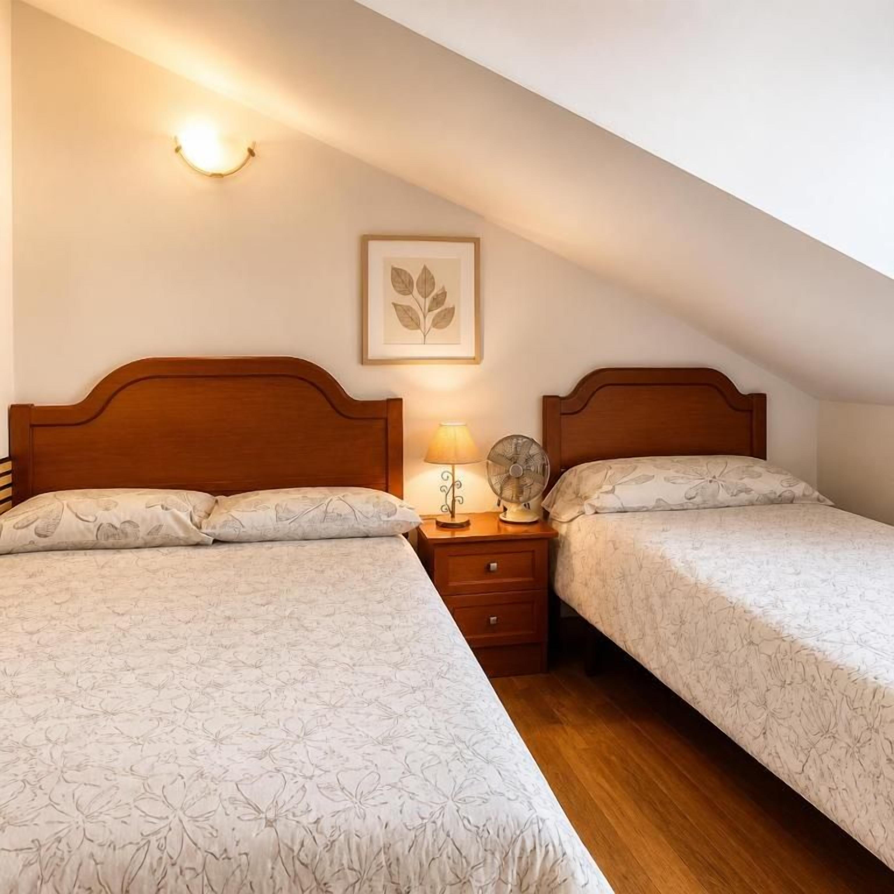 A bedroom with two beds, a bedside table with a lamp, a wall-mounted fan, and a framed picture.