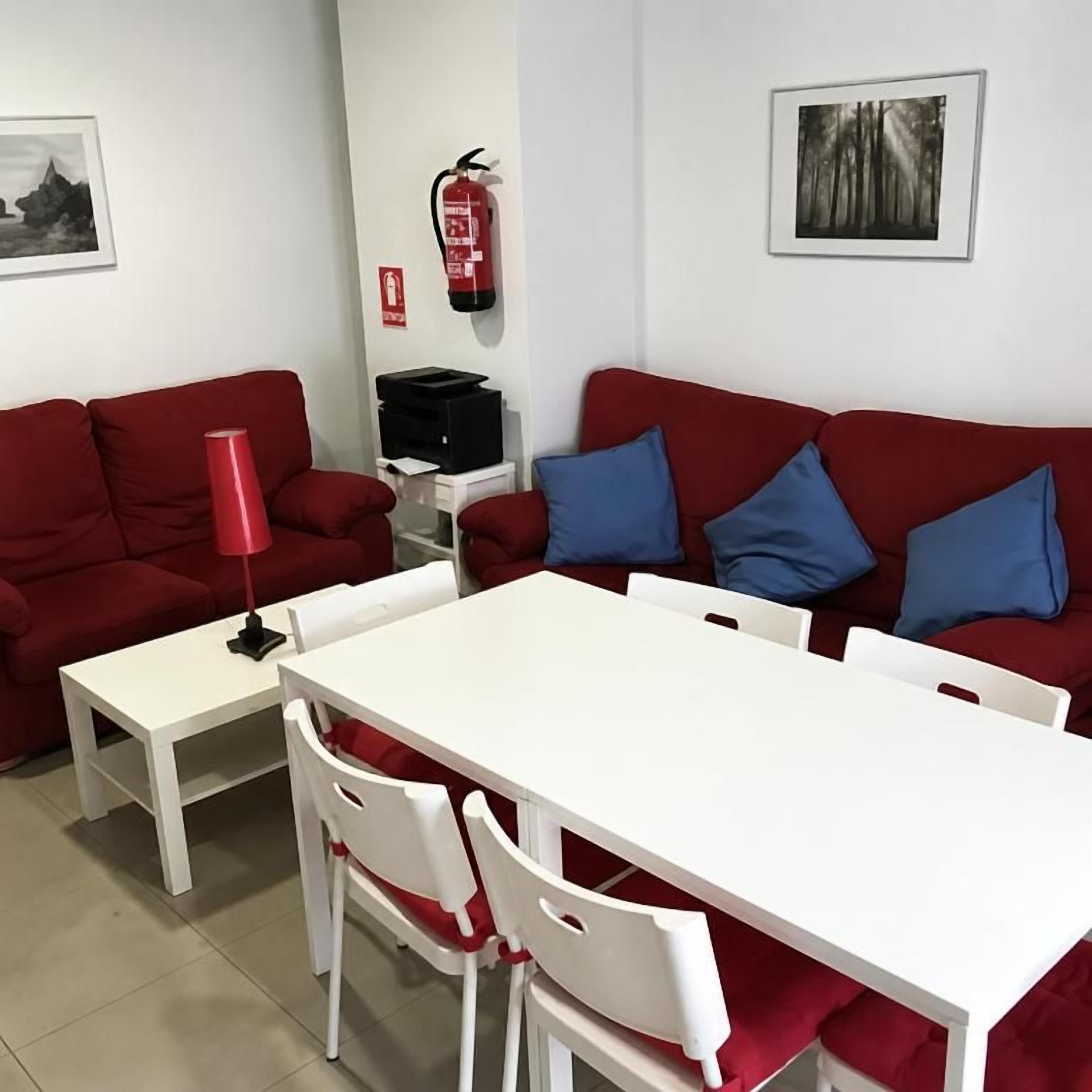 A room with a red sofa, white table, and chairs, a fire extinguisher, and framed pictures on the walls.