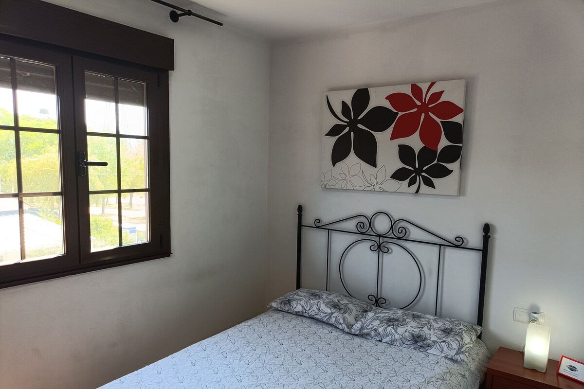 A bedroom with a single bed, a nightstand, a wall-mounted lamp, and a window with a view of greenery.