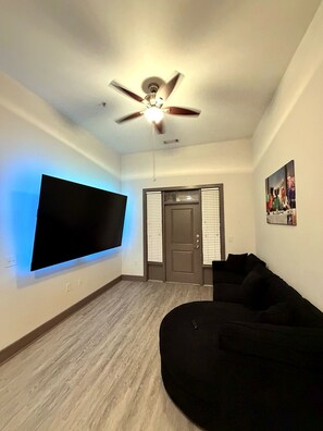 Ein Wohnzimmer mit einem Wand-Fernseher, einem Deckenventilator, einer Tür mit Jalousien und einem schwarzen Sofa.