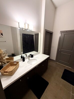 Un baño con tocador blanco, un espejo grande, una puerta oscura y piso de baldosas.