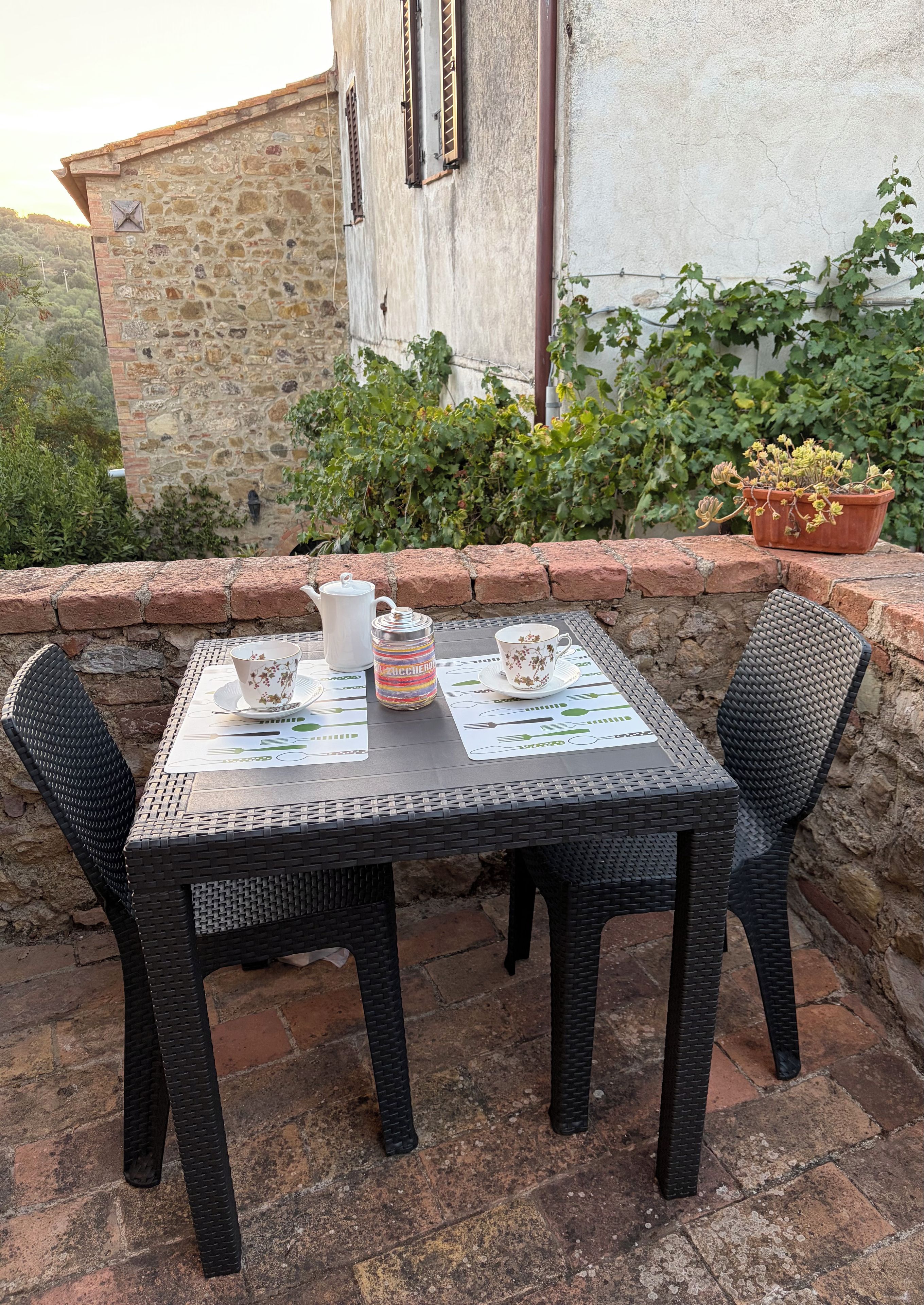 Une table dressée à l’extérieur, avec des chaises en osier, une théière blanche, une tasse et une bouteille.