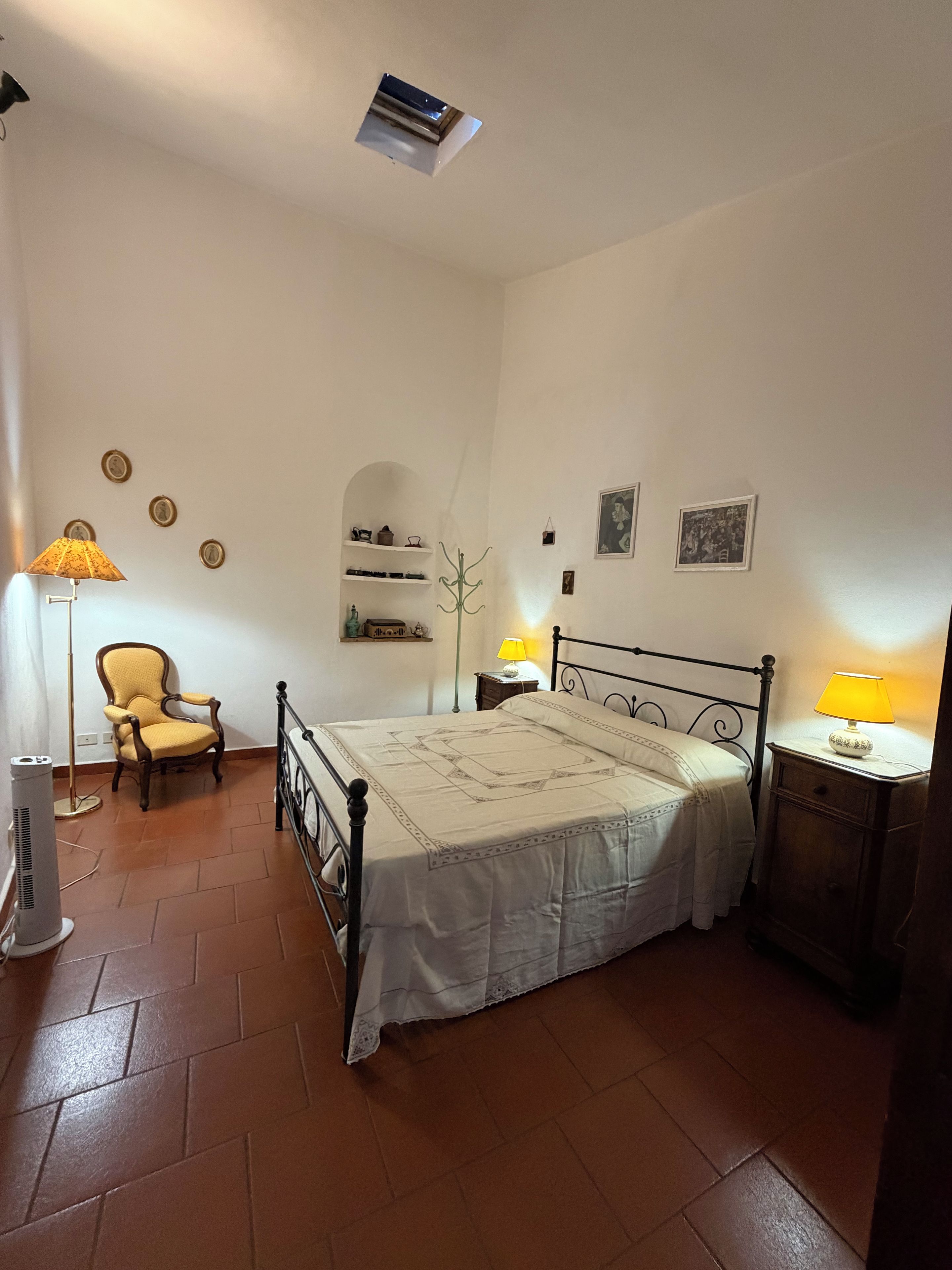 A bedroom with a bed, a chair, a lamp, a heater, and a shelf with decorative items.