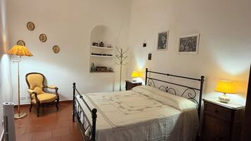 A bedroom with a bed, a chair, a lamp, a heater, and a shelf with decorative items.
