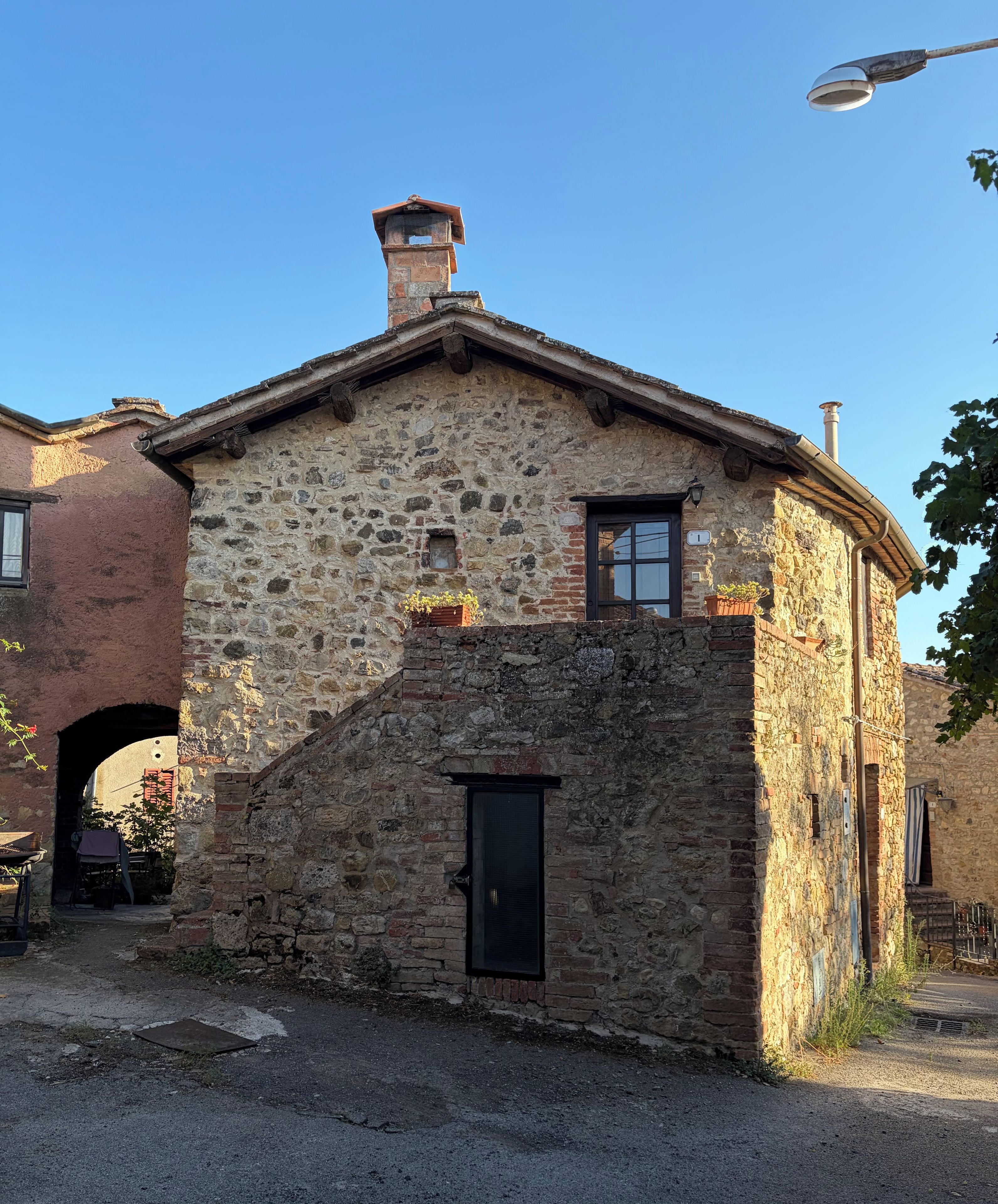 Ein Steinhaus mit kleinem Fenster und einer Tür, umgeben von einer gepflasterten Fläche und einem Backsteinbogen.
