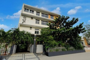 A multi-story residential building with a gated entrance, surrounded by lush greenery and a clear blue sky.