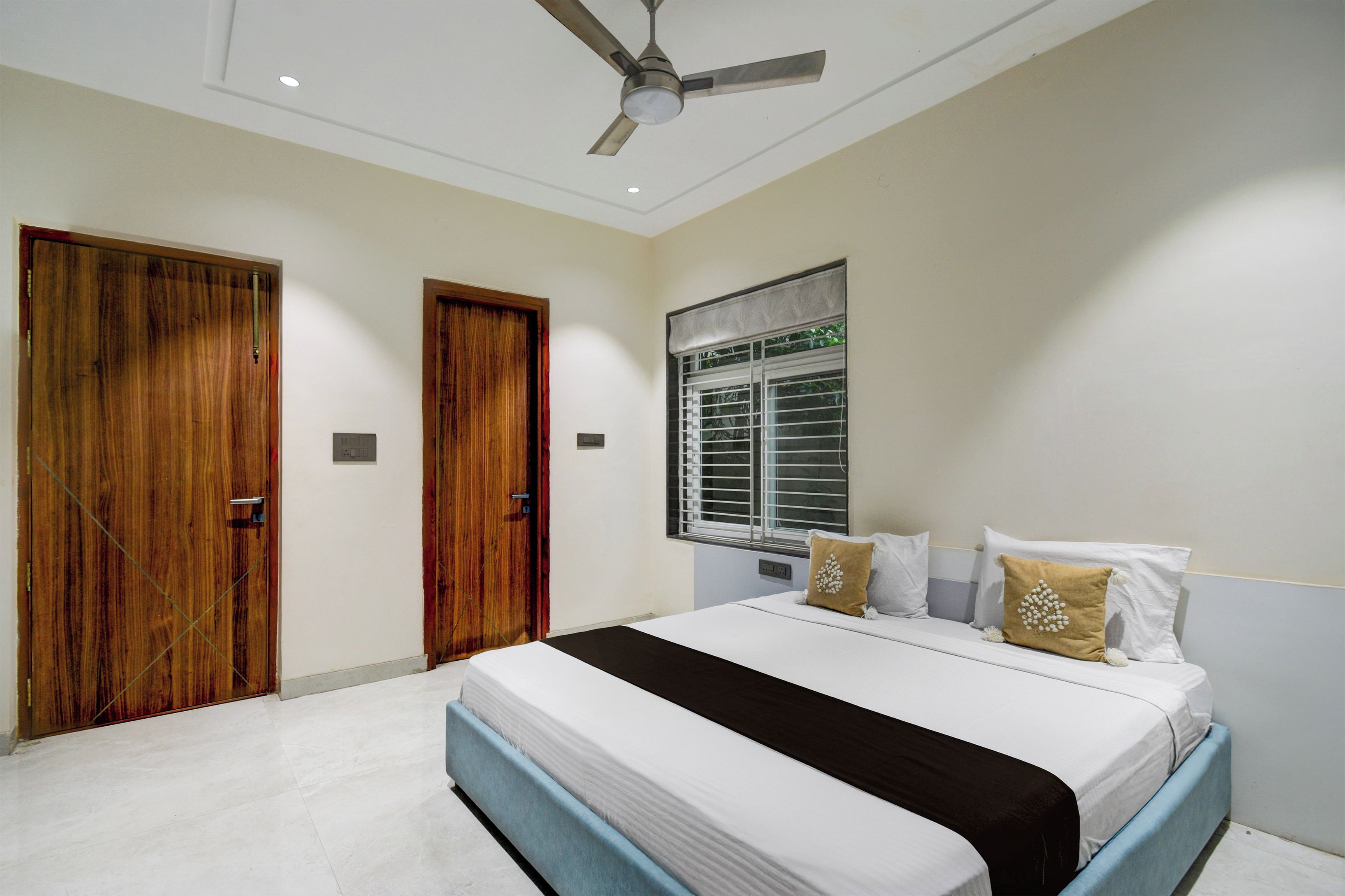 A bedroom with a bed, two wooden doors, a ceiling fan, a window with blinds, and a wall-mounted light fixture.