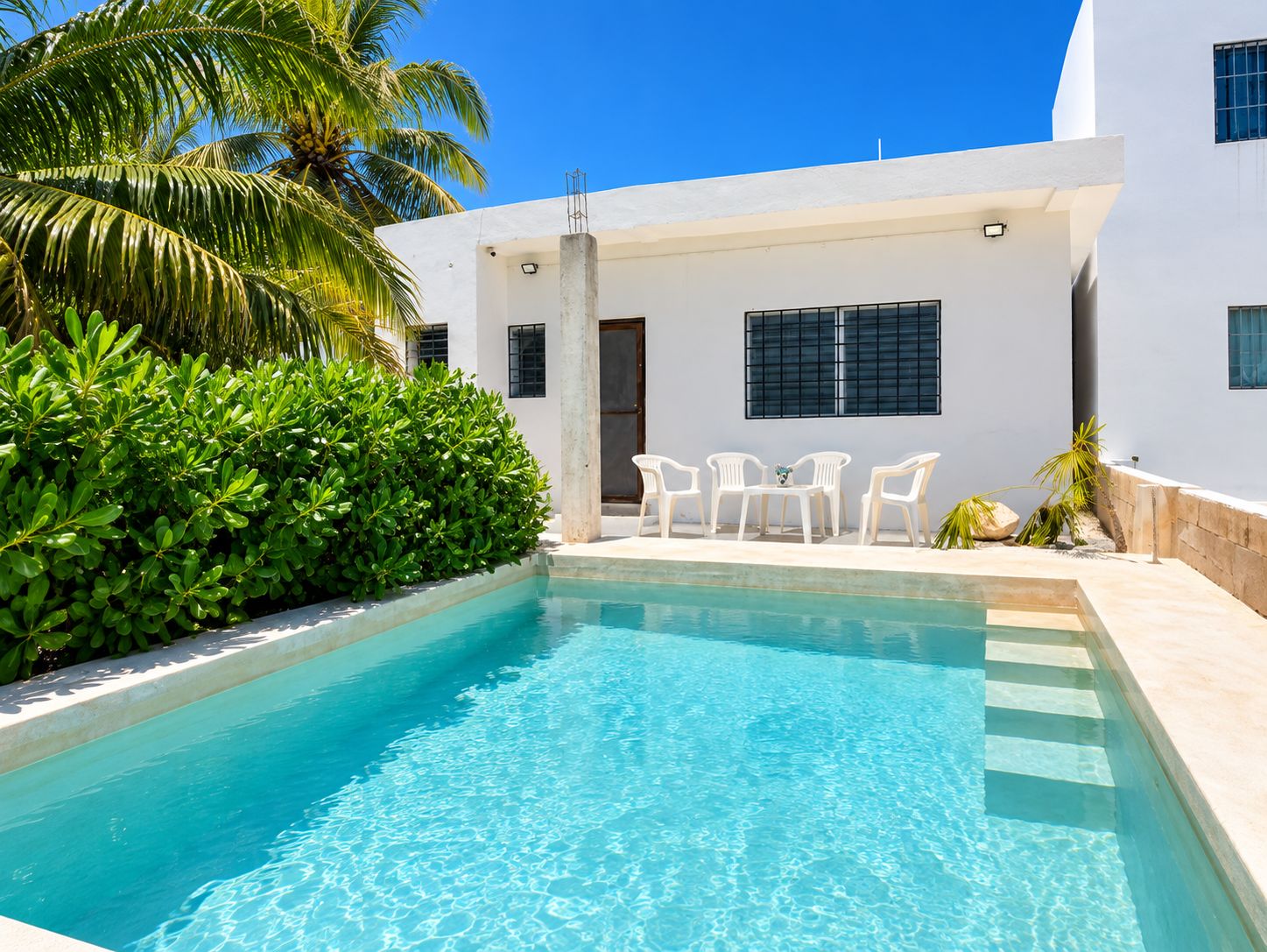 A swimming pool with clear blue water, surrounded by white walls and a palm tree. There are white plastic chairs and a small table by the pool.