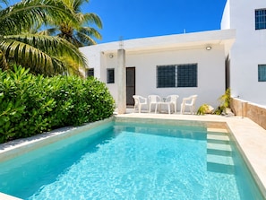 A swimming pool with clear blue water, surrounded by white walls and a palm tree. There are white plastic chairs and a small table by the pool. - Mar-e-Luna | Private Pool & Beach Living in Chicxulub (Chicxulub)
