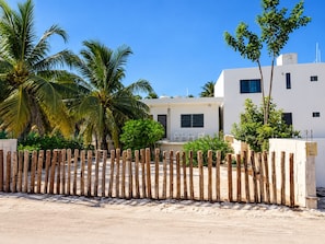 A white building with a sandy ground, surrounded by a wooden fence, palm trees, and a clear blue sky. - Mar-e-Luna | Private Pool & Beach Living in Chicxulub (Chicxulub)