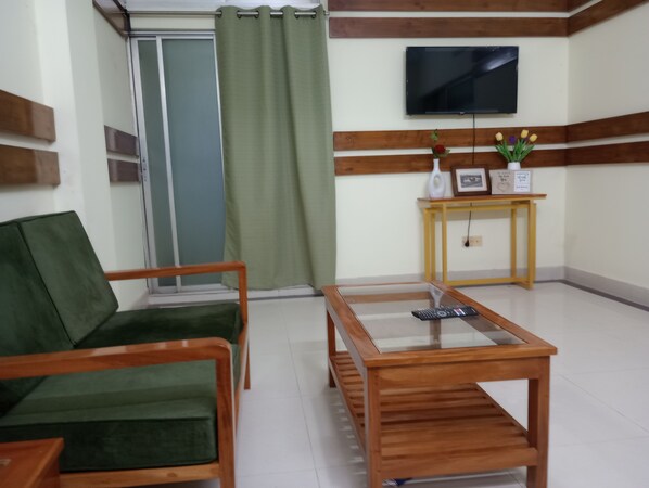 A room with a green sofa, a wooden coffee table, a television mounted on the wall, and a small table with a vase and picture frame.