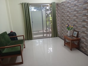 Ein Zimmer mit einem grünen Sofa, einem Holztischchen mit einer Blumenvase und einer Glastür, die auf einen Balkon führt.