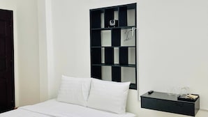 Ett hotellrum med en säng, en svart förvaringsenhet, en papperskorg och en vägghylla med glas och en kamera.