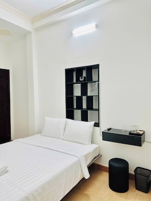Une chambre d’hôtel avec un lit, un meuble de rangement noir, une poubelle et une étagère fixée au mur, sur laquelle se trouvent des verres et un appareil photo.