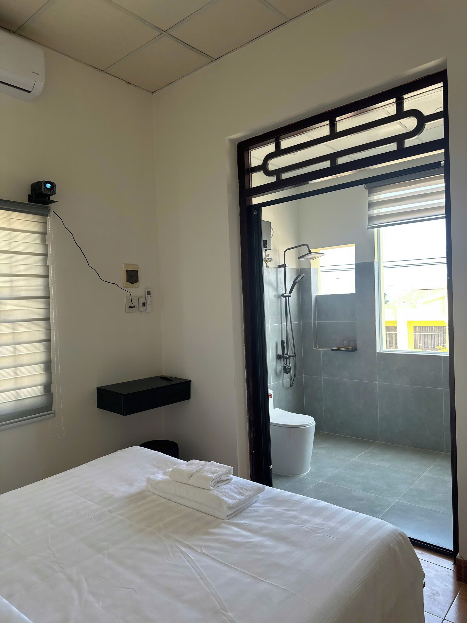 Une chambre d’hôtel comprenant un lit, une salle de bain avec douche, une fenêtre avec stores et un climatiseur fixé au mur.