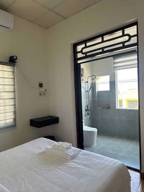 Une chambre d’hôtel comprenant un lit, une salle de bain avec douche, une fenêtre avec stores et un climatiseur fixé au mur.