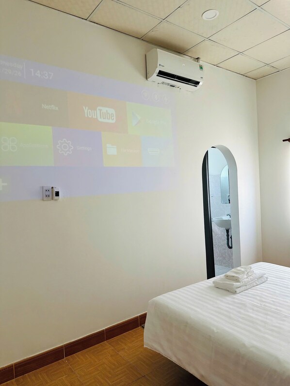 A hotel room with a bed, a wall-mounted air conditioner, a projection screen displaying app icons, and a wooden floor.