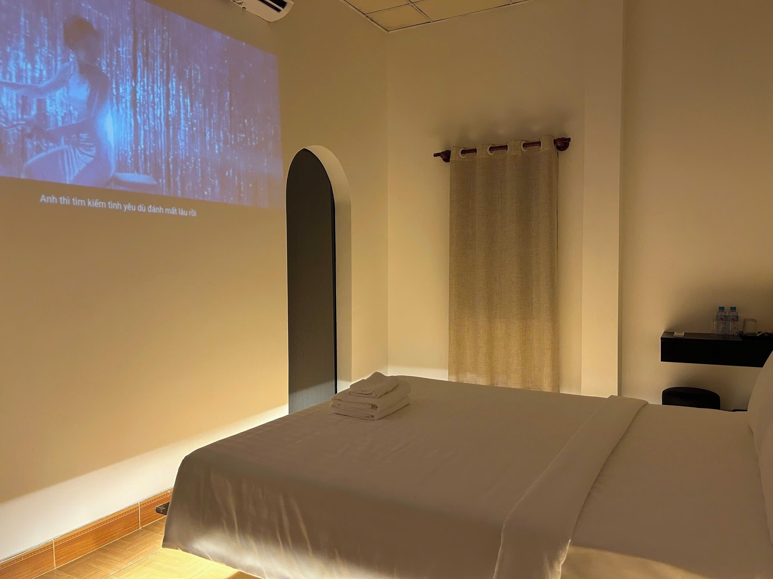 Une chambre d’hôtel équipée d’un lit, d’un écran de projection, d’un rideau et d’une étagère avec des bouteilles d’eau.