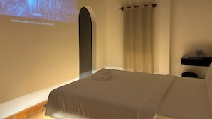 Ett hotellrum med en säng, en projektionsduk, ett draperi och en hylla med vattenflaskor.