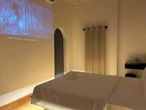 Une chambre d’hôtel équipée d’un lit, d’un écran de projection, d’un rideau et d’une étagère avec des bouteilles d’eau.