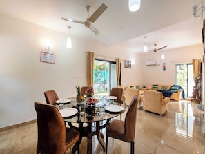 A dining area with a round table, chairs, and a fruit bowl. There is a sofa and a window with curtains in the background. - DanCenter Trizi Villa 4BHK with Pvt Pool (Lonavala)
