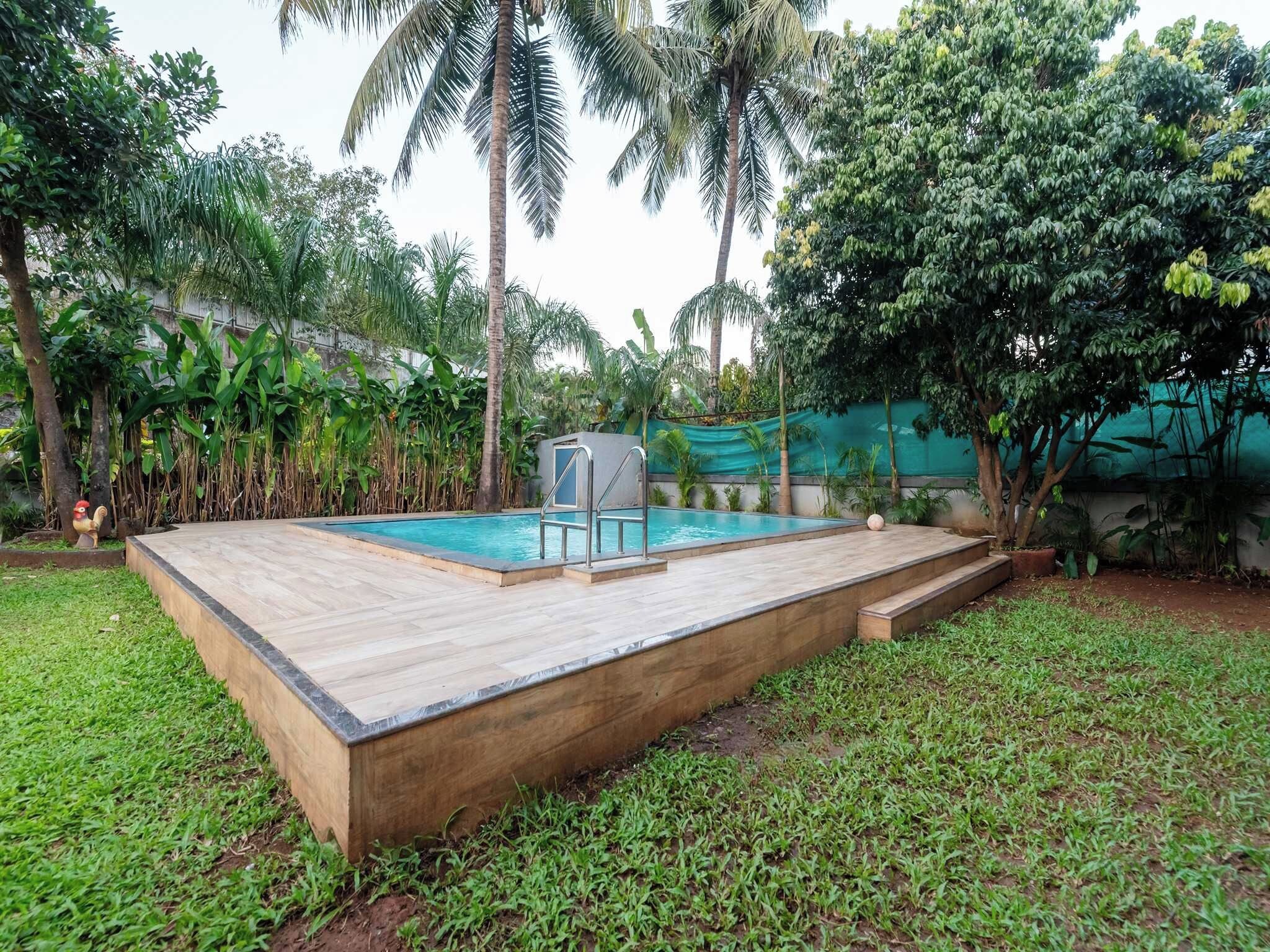 A rectangular swimming pool with a wooden deck, surrounded by lush greenery and palm trees.