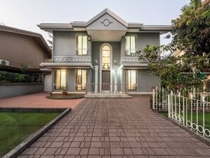 A two-story house with a symmetrical facade, a central entrance with a decorative arch, and a paved driveway leading to the front door. - DanCenter Trizi Villa 4BHK with Pvt Pool (Lonavala)