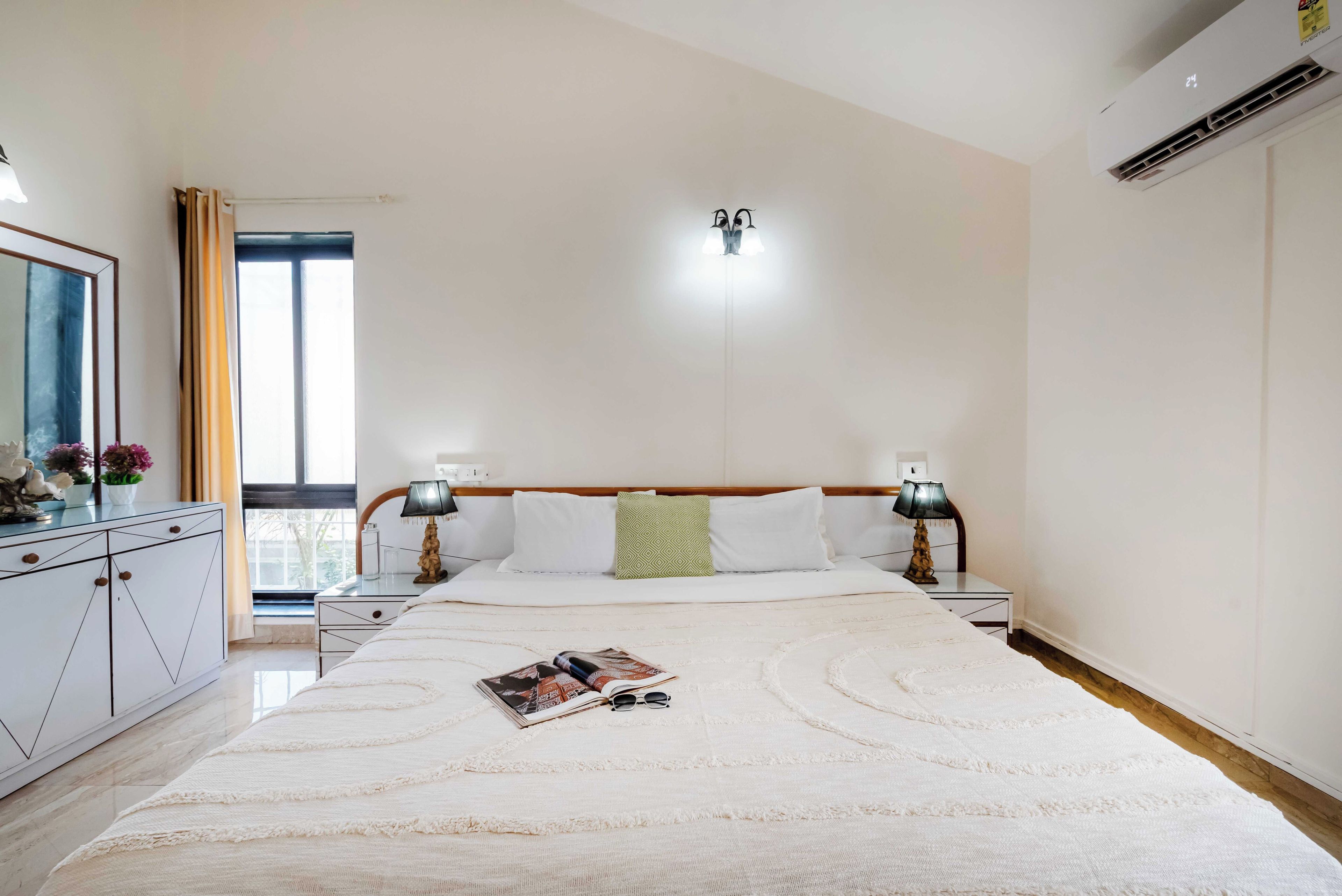 Ein Hotelzimmer mit Bett, Nachttischen, einem Spiegel, einem Fenster mit Vorhängen und einer Klimaanlage.