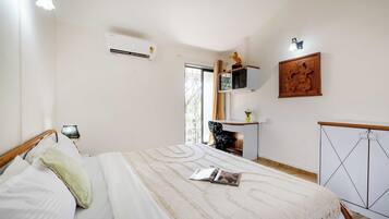 A bedroom with a bed, a bedside table, a chair, an air conditioning unit, a window with curtains, and a wall-mounted shelf with decorative items.