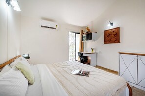 A bedroom with a bed, a bedside table, a chair, an air conditioning unit, a window with curtains, and a wall-mounted shelf with decorative items. - DanCenter Trizi Villa 4BHK with Pvt Pool (Lonavala)