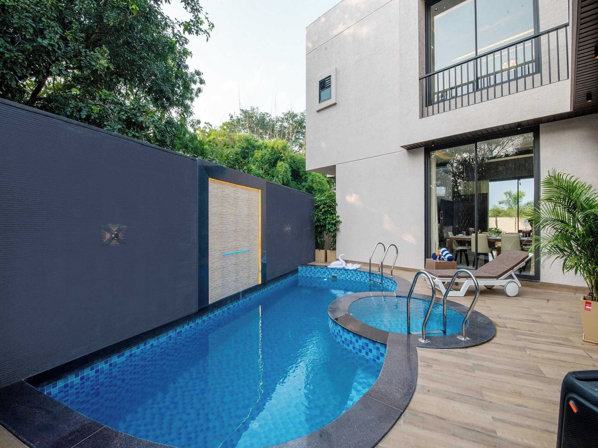 A swimming pool with a tiled edge, a lounge area with chairs, and a building with a balcony.