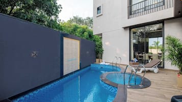 A swimming pool with a tiled edge, a lounge area with chairs, and a building with a balcony.