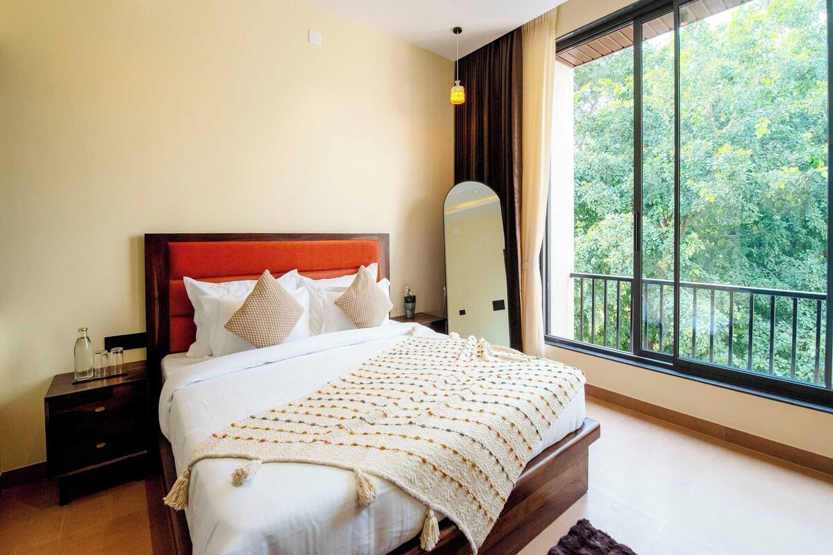 Quarto com cama grande, mesa de cabeceira, espelho e janela com vista para o verde.