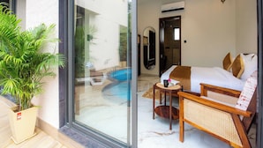 Quarto de hotel moderno com porta de vidro para uma piscina, ventilador de teto e pequena mesa com um livro e uma xícara.