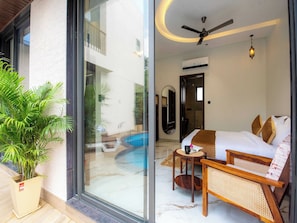 A modern hotel room with a glass door leading to a pool, a ceiling fan, and a small table with a book and a cup. - DanCenter Papillon Villa 5BHK with Pool (Udaipur)