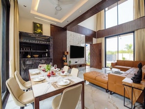 A modern living room with a dining area, a flat-screen TV, and a large window offering a view of the outdoors. - DanCenter Papillon Villa 5BHK with Pool (Udaipur)