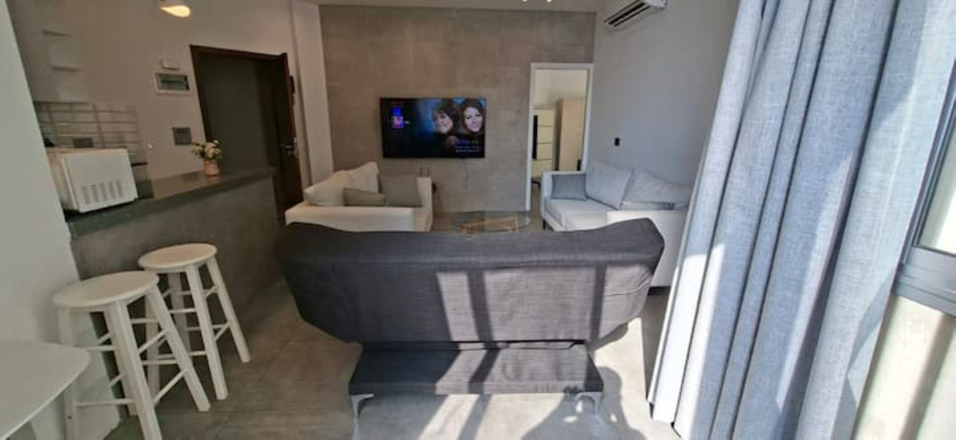 A modern living room with a sofa, two white bar stools, a flat-screen TV mounted on the wall, and a window with curtains.