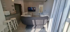 Ein modernes Wohnzimmer mit einer Couch, zwei weißen Barhocker, einem wandmontierten Flachbildfernseher und einem Fenster mit Vorhängen.
